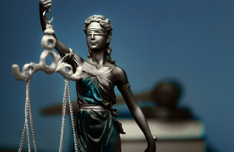 Figura Temidy z opaską na oczach i wagą w dłoni, symbolizująca sprawiedliwość, na tle młotka sędziowskiego i książek prawniczych.