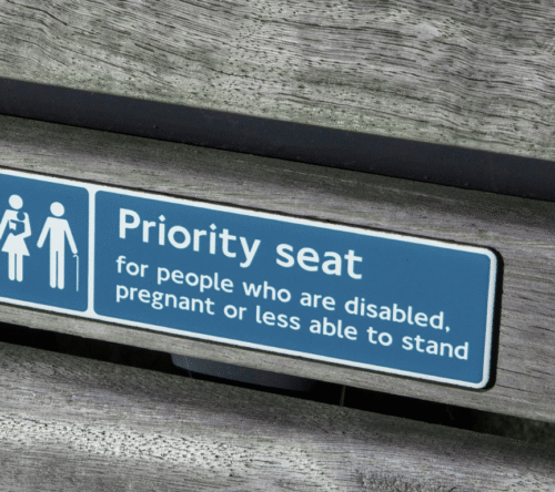 Tabliczka informacyjna z napisem „Priority seat for people who are disabled, pregnant or less able to stand”, umieszczona na drewnianej ławce – symbolicznie przedstawia uprzywilejowane miejsce siedzące dla osób z niepełnosprawnościami, kobiet w ciąży oraz osób z ograniczoną mobilnością.