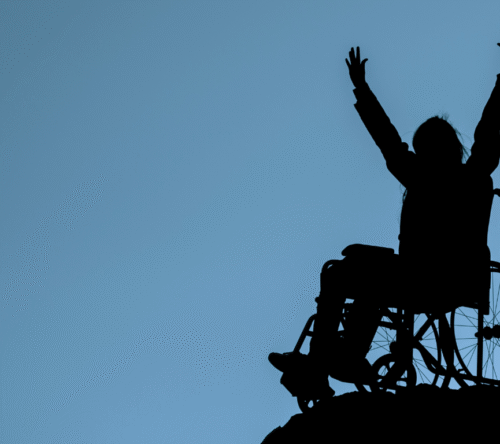 Osoba na wózku inwalidzkim z uniesionymi rękami na tle nieba – symbol determinacji i przełamywania ograniczeń.